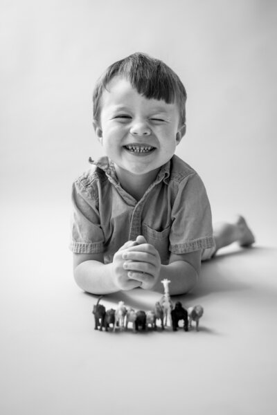 Black and white photo of young boy laying on floor winking at camera and smiling while a line of small toy animals is in front of him at a studio in Annapolis Maryland
