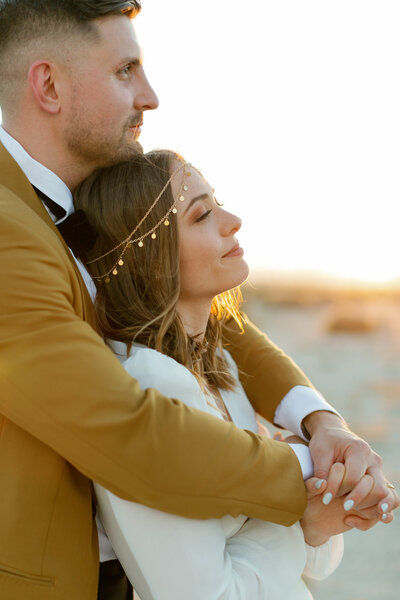 groom holding his bride from behind and clasping hands in the front. both are looking off with a soft smile at the sunset.