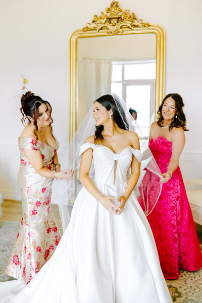 Bride in an off-the-shoulder satin gown smiles while her mother and mother in law adjust her veil during a joyful spring wedding morning in North Texas.

