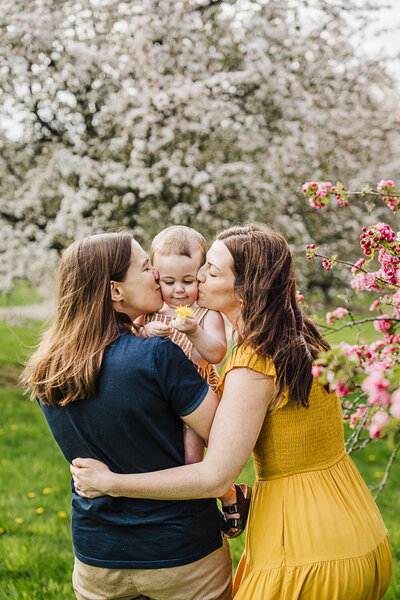 two mothers kiss baby boy on cheeks in spring blossoms in boston