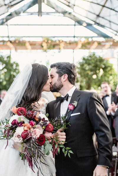 Bride and groom share a romantic first kiss under the greenhouse canopy at Planterra Conservatory, surrounded by lush greenery, string lights, and emotional guests during a black tie wedding ceremony.