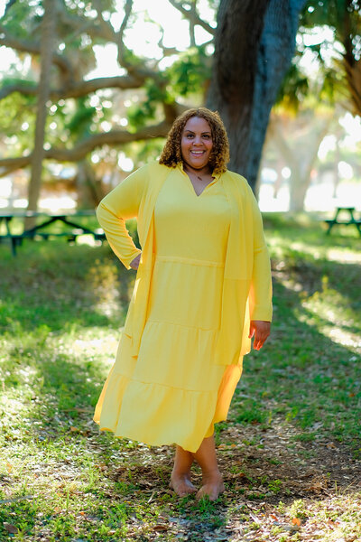 Certified life coach Claudine Martinell stands barefoot in a vibrant yellow dress, ready to help you explore the landscapes of your life.