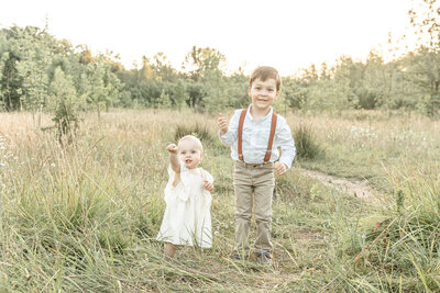 One year old girl in a cream dress  standing out in nature with older  brother dressed in khaki pants, white shirt and brown suspenders looking at camera during their sw portland oregon family photoshoot
