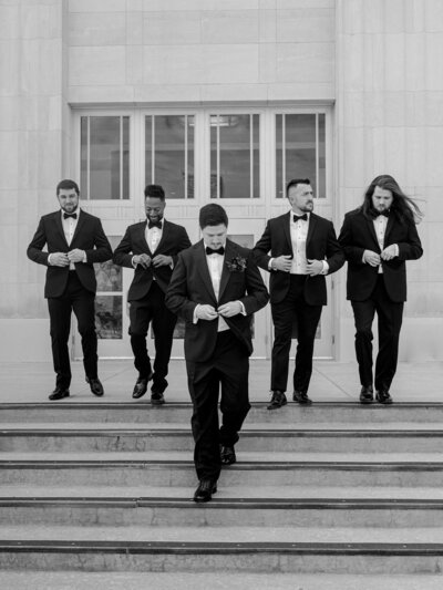 A groom and his groomsmen walk down the steps of a wedding venue in an editorial portrait.