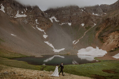 Couple in wedding attire walk in front of alpine lake in the San Juan Mountains