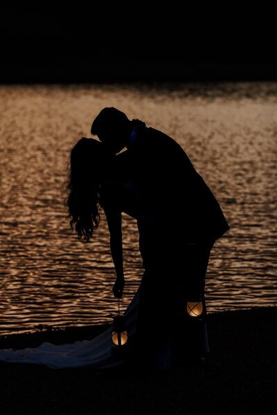 Silhouette of bride and groom against lake at sunset during intimate wedding.