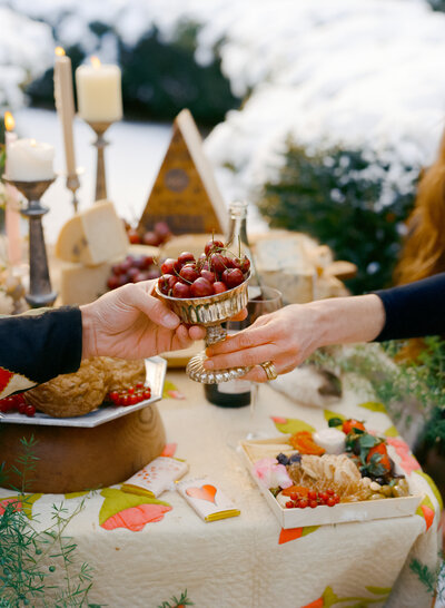 A warm and inviting 35mm film photograph from a Valentine's Day branding session for a fine foods store and florist collaboration. The image captures a heartfelt moment as two hands exchange a silver goblet filled with cherries, surrounded by a beautifully styled table. The spread includes artisanal cheeses, fresh fruits, candles, and delicate floral touches, all set against a snowy outdoor backdrop. This cozy, romantic scene perfectly showcases the collaboration's thoughtful and elegant offerings for celebrating love.