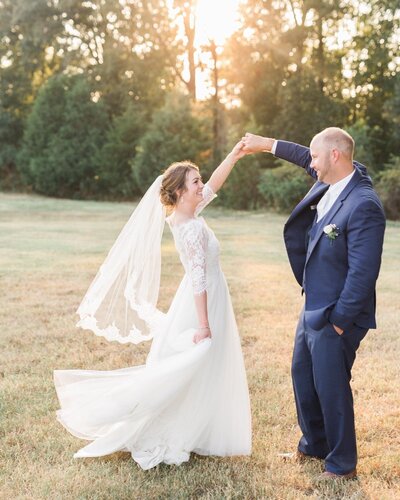 Bride in dress with sleeves and with veil dancing with groom in navy suit during golden hour for newlywed pictures in field