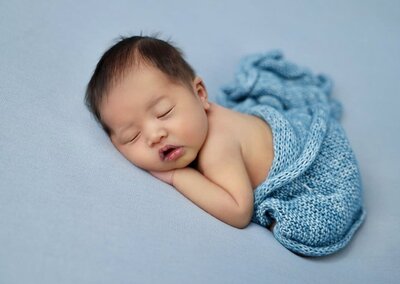 baby boy lying on stomach wrapped in a blue blanket and laying against a blue backdrop.