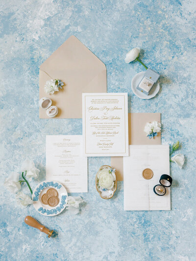 Flay lay of a white and blue themed wedding invitation suite and wedding rings surrounded by pink flowers and rings on a blue and white background