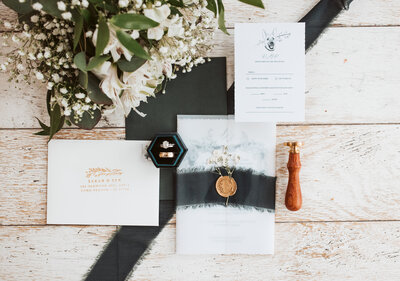 Wedding invitation suite with rings, wax seal, and bouquet styled in a flat lay on rustic wood for a Maine wedding.