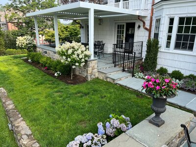 A beautifully transformed home entrance featuring a lush green lawn, vibrant flower beds, a stone pathway, and a charming covered porch with outdoor seating.