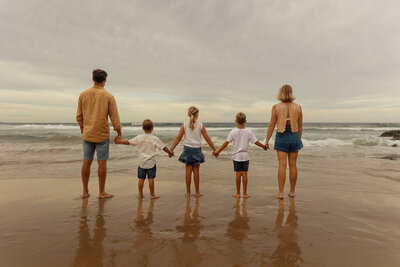 A family portrait of them holding hands on a Sunshine Coast beach looking out to the ocean. Photo taken from behind