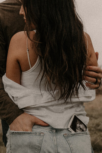 A couple hold each other while the husband puts his hand in his wife's pocket and a sonogram print peeks out of the other pocket. 