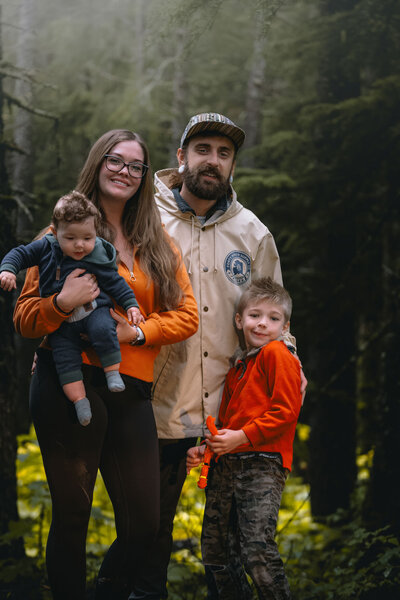 A family of four standing together in a forest, with the woman holding a baby and the man standing beside a young boy. They are dressed warmly, surrounded by tall green trees.