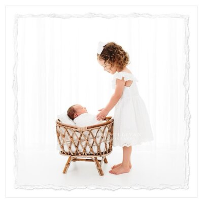 The photo shows a sweet moment between a newborn baby and her older sister. The newborn is peacefully lying in a wicker bassinet, while her big sister tenderly touches her. Both girls wear all white and the background is all white, creating a serene and ethereal atmosphere. This captivating image was captured by photographer Bri Sullivan.