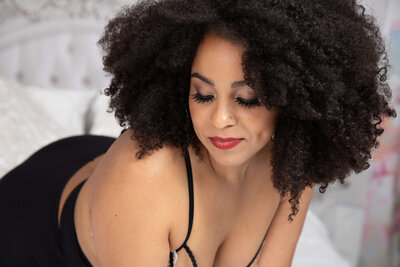 A woman in red lingerie is lying on her back on a white ornate bed with her legs raised and crossed. She has long hair, and her arm is resting on her forehead. The background features a white textured wall and decorative pillows.