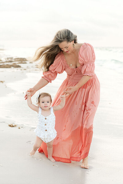 A mom in a coral dress helps her daughter take sandy steps along the beach, capturing a joyful and windy sunset moment.