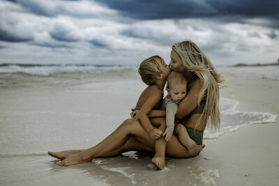 Family Photographer, a  has both kids close to her in her lap, a young son and a baby, at the ocean
