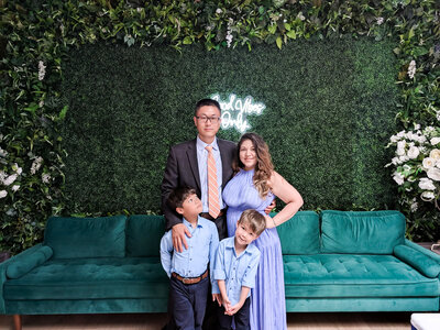 Teresa's husband, and two young sons are dressed in formal attire, smiling at the camera, in front of a green couch and grassy decorative backdrop wall.