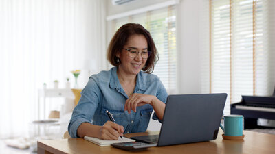 A Filipina woman smiles as she takes notes from a virtual meeting with her laptop in front of her