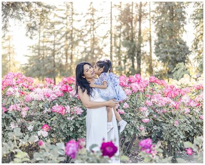 Toddler daughter kissing her mother amid blooming roses in San Jose rose garden during a family photoshoot