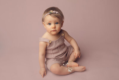 a pretty one year old girl wearing a purple knitted romper holding flowers on a purple backdrop in Rochester, NY