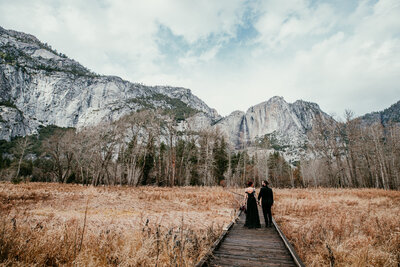 Couple dressed in black walking hand in hand in Yosemite National Park, with Half Dome towering in the background, featured in a 'How to Plan Your Yosemite Elopement' guide.