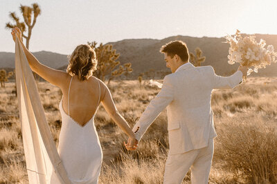 Bride and groom holding hands, walking through the golden desert landscape of Joshua Tree National Park at sunset.
