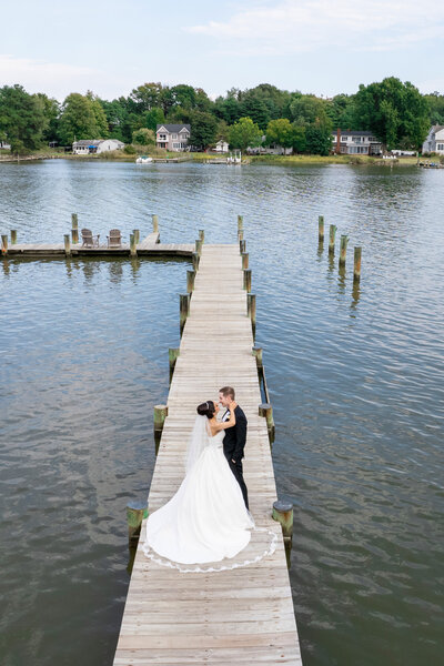 A bride and groom stand on a wooden dock over a serene lake. The bride wears a flowing white gown and veil, and the groom is in a black suit. They embrace near the end of the dock, surrounded by calm water and lush greenery in the background.