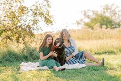 Mom in striped dress and daughter in green dress on a blanket in a field with their chocolate lab dog
