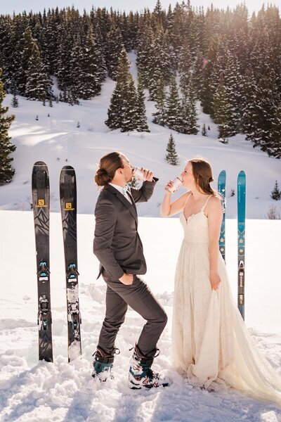 A couple in wedding attire drink hard seltzers next to their skis in the snow in Colorado