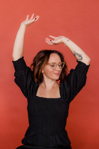 Woman poses for the camera against a colored backdrop with eyes closed and both hands in the air.