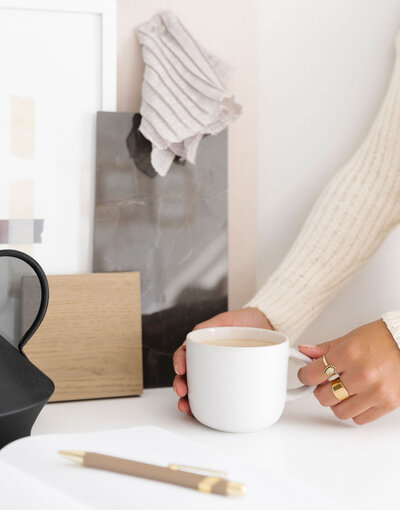 white wall, neutral material samples leaned on wall, brown and gold pen on desk, woman's hands with gold rings holding a white mug of coffee