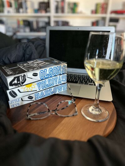 Stack of Blue Devils Hockey books by S.J. Sylvis with custom sprayed edges from Sprayed Your Pages, styled on a desk with a glass of wine.