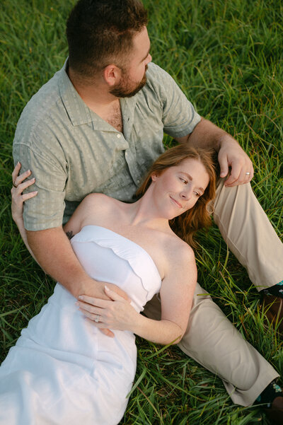 In the midst of a romantic picnic in the park, a couple sits on a white blanket. Beside them, a woven basket holds a collection of wildflowers. The girl turns towards the camera, as the guy captures the enchanting scene with his photography.