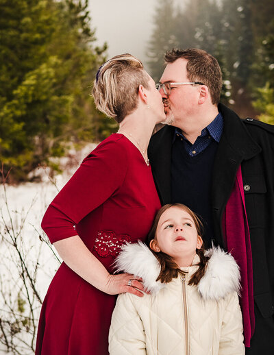 Parents kiss while their daughter expresses discomfort at their family photoshoot session in the mountains north of Seattle WA.