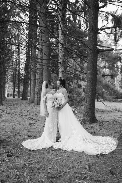 LGBTQ couple in their wedding dresses looking at each other and embracing in a woods setting.
