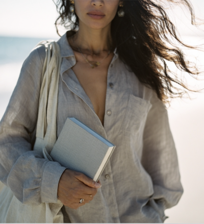 A woman holding a linen notebook on the beach wearing a neutral shirt, symbolizing the creative freedom and intentional lifestyle behind Kathalyst Design Studio.
