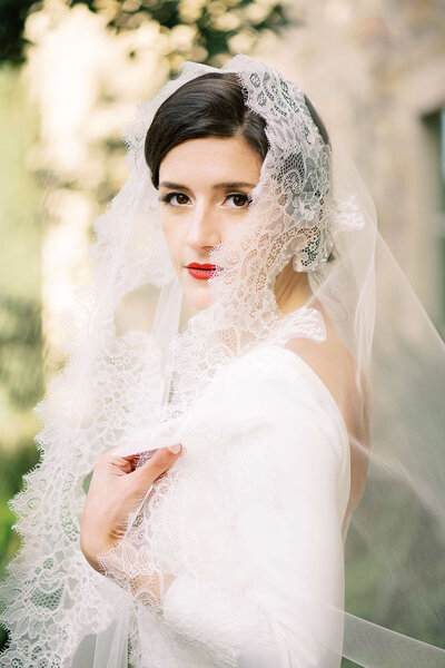 Classy bride in long sleeved wedding dress with red lipstick looks into the distance while holding bouquet