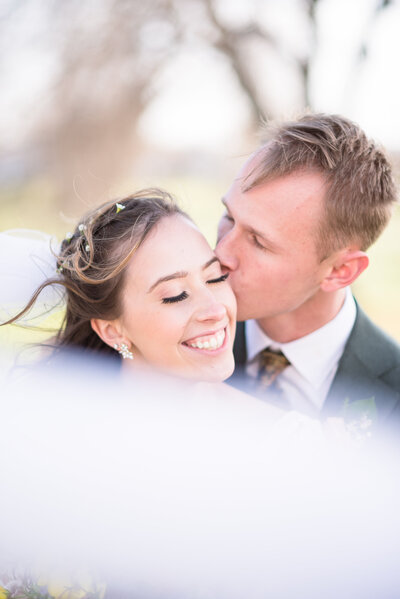 groom holding his bride around the waist and kissing her forehead as the bride smiles over her shoulder at him captured by colorado wedding photographer