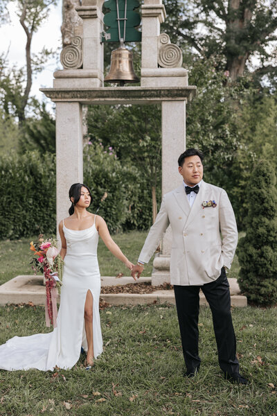 Bride and groom holding hands beneath an elegant stone bell structure during their outdoor wedding portraits, captured by a documentary-style Virginia wedding photographer.