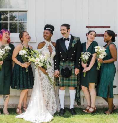 Bride and groom in a white wedding gown and kilt standing with bridesmaids.
