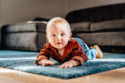 A baby crawling on a blue rug in a well-lit living room, looking directly at the camera with a curious expression.