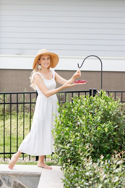 A woman in a garden hanging a feeder of homemade hummingbird food.
