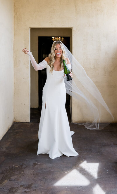 Joyful bride laughing as she pops a bottle of champagne to celebrate her wedding day at Ma Maison