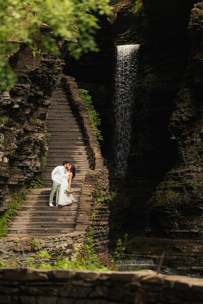 a newlywed couple kissing on a stone staircase overlooking a waterfall in seneca lake new york