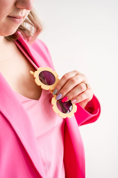 Kat Murphy holding orange flower sunglasses and wearing a hot pink blazer