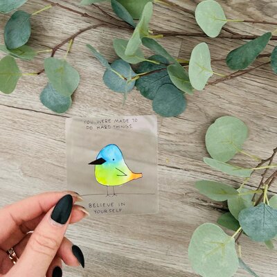 A hand with black fingernails holding a little watercolor painted bird.