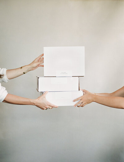 hands give stack of white branded boxes, packaging to other set of hands, clean background, film branding photographer in Michigan